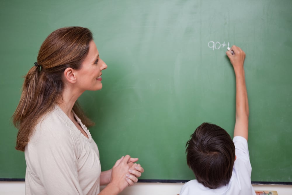 Happy schoolteacher helping a schoolboy doing an addition on a blackboard