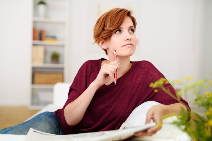 Thoughtful Attractive Young Woman Answering Crossword Puzzle Game on Newspaper at the Living Room Couch.