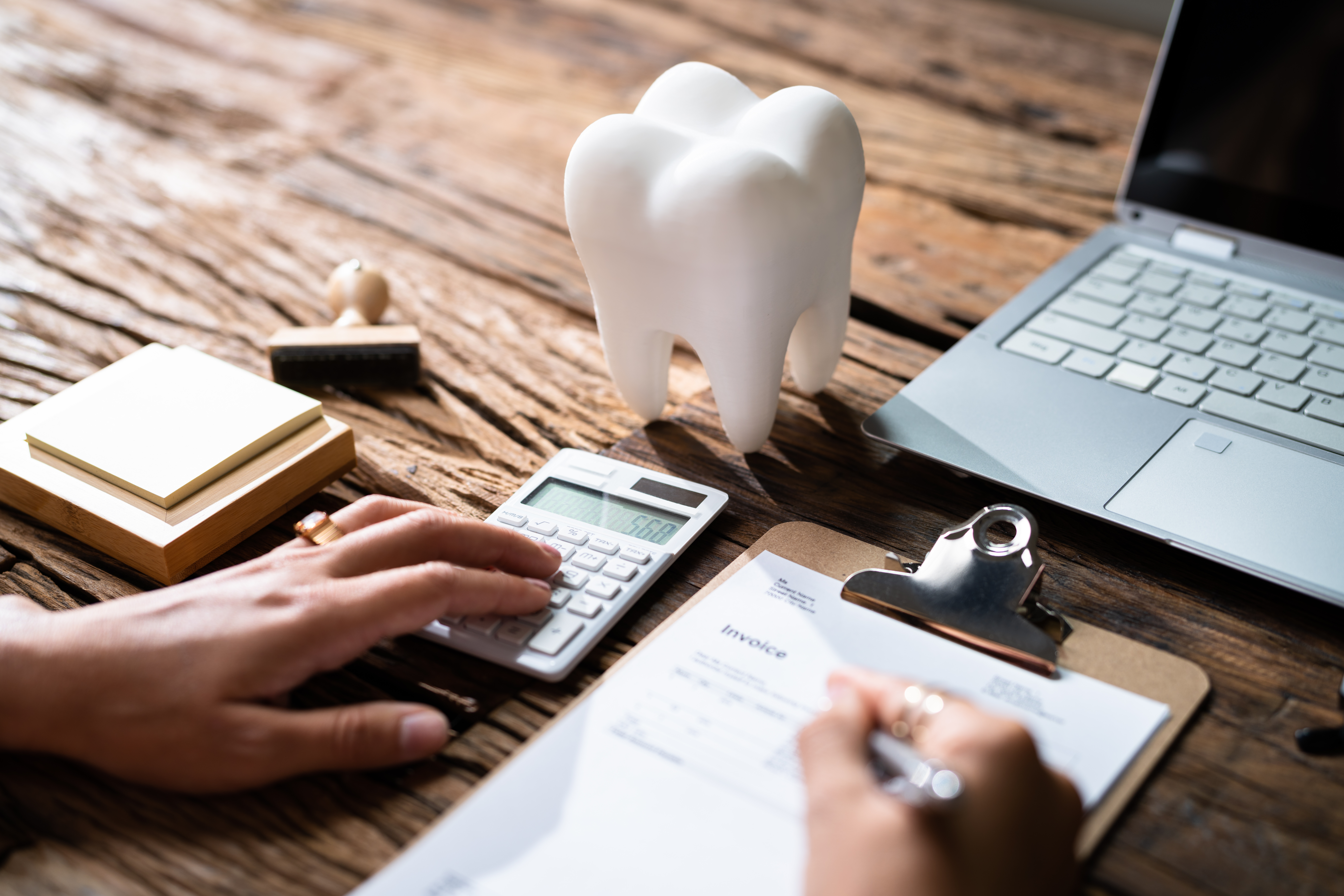 A set of hands on a table using a calculator and writing on a clipboard with a large tooth display in front of them comparing dental plan pricing models